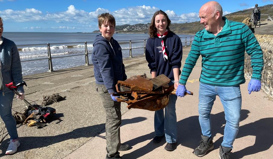 Picture taken beside the beach with 3 people carrying some rusty rubbish wearing gloves